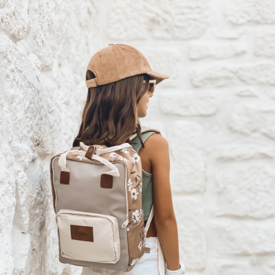 Mochila bege com flores brancas usada por criança com boné e óculos junto a parede branca