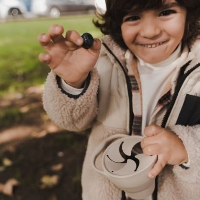 Criança com casaco bege segurando uma bola azul e um frasco transparente ao ar livre