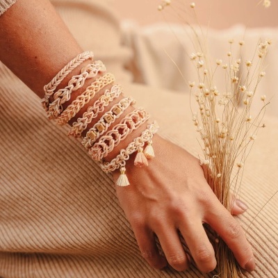 Pulseiras trançadas bege e rosa com detalhes dourados e pérolas, com franjas, no pulso segurando flores secas.