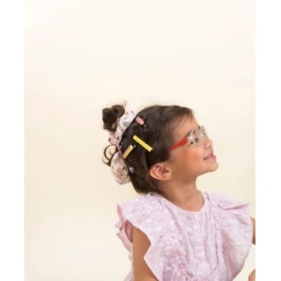 Menina com óculos vermelhos, cabelo com acessórios e blusa rosa em fundo neutro