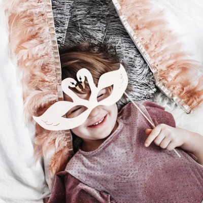 Criança com máscara branca de cisne e camisola rosa de veludo deitada sobre tecidos brancos e rosa claro.