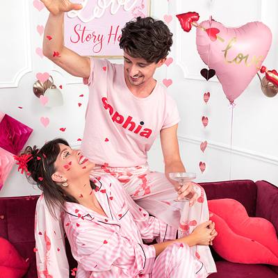 Casal com pijamas rosa sentados num sofá vermelho com decoração de balões cor de rosa em formato de coração