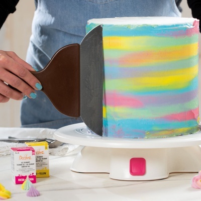 Bolo colorido em tons pastel a ser alisado com espátula e caixa de corantes Decora