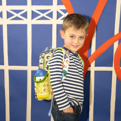 Criança com mochila infantil colorida e garrafa azul no bolso lateral num fundo azul com padrões laranja e branco