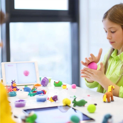 Criança a moldar massinha rosa numa mesa com várias figuras de massinha coloridas e tablets