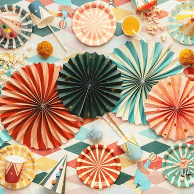 Decorações de festa coloridas em papel com leques, chapéus, pratos e copos sobre mesa com toalha estampada