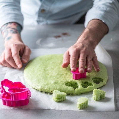 Mãos cortando massa verde com cortador de biscoitos rosa em papel vegetal