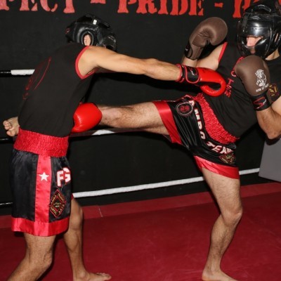 Dois homens a treinar Muay Thai em ringue, com equipamento de proteção e calções pretos e vermelhos