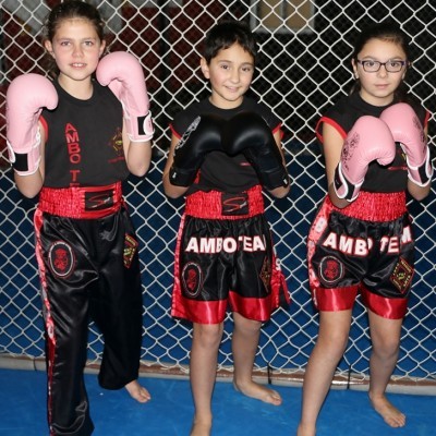 Três crianças com roupa de boxe preta e vermelha e luvas rosas e pretas em piso azul com rede metálica ao fundo