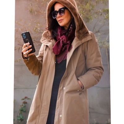 Mulher com casaco castanho claro com capuz de pelo, lenço bordeaux, óculos pretos e telemóvel com capa AMG