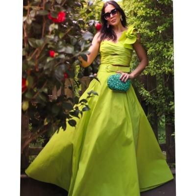 mulher com vestido verde limão longo, bolsa azul esverdeada, óculos de sol, rodeada de plantas e flores vermelhas