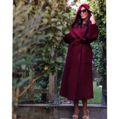 Mulher com casaco bordô comprido com capuz e cinto, usando óculos de sol, em jardim com plantas ao redor