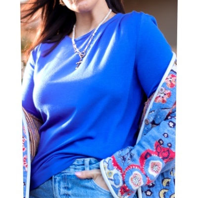 Mulher com blusa azul e casaco colorido com padrão, jeans azul claro, colares e óculos de sol.