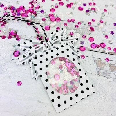 Embalagem decorativa branca com bolinhas pretas e laço, com janela oval mostrando lantejoulas rosa, cordão branco e preto, sobre fundo branco com lantejoulas espalhadas