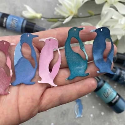 Cinco pins coloridos em forma de pinguim numa mão com flores e frascos de tinta ao fundo