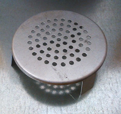 Tampa redonda perfurada em metal prateado sobre superfície metálica cinza