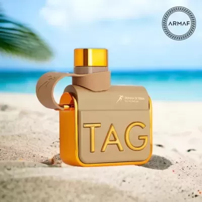 Frasco de perfume TAG bege e dourado na areia da praia com mar azul ao fundo