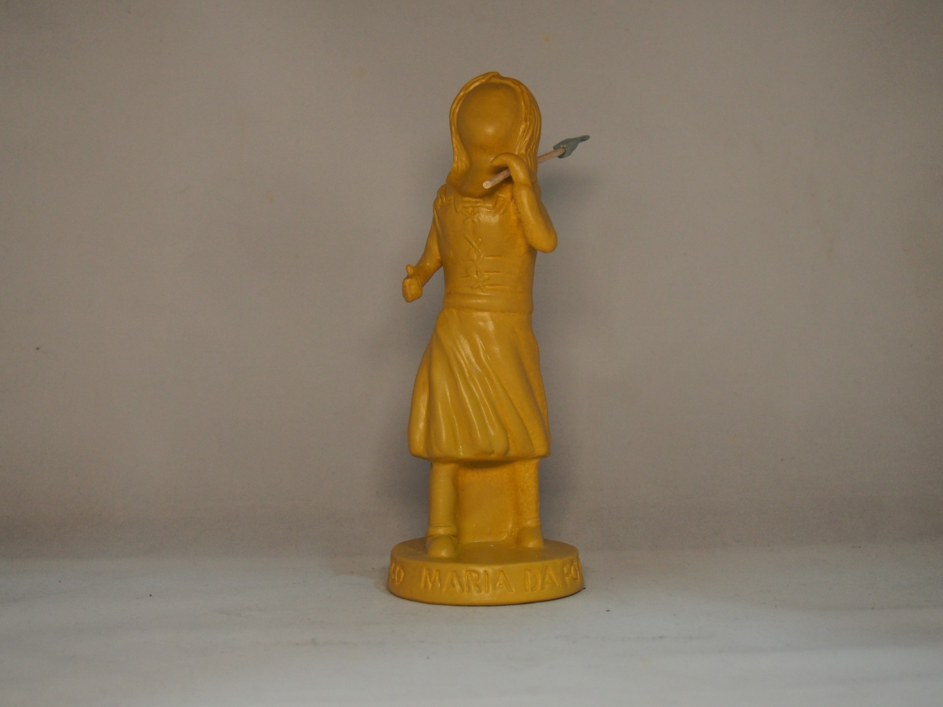 Estatueta amarela de menina com vestido e chapéu em fundo cinza