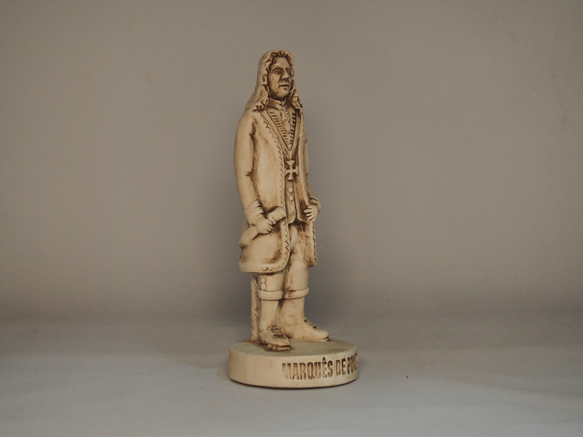 Estátua pequena de homem vestido com roupa antiga em tom bege claro sobre base redonda