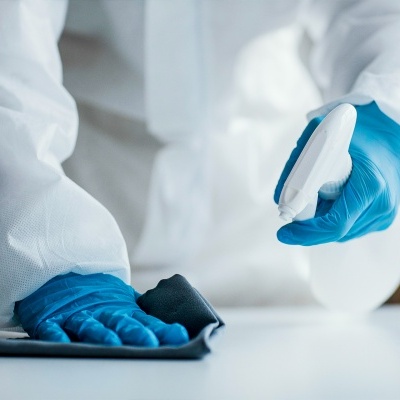 Pessoa a limpar superfície com pano e borrifador, luvas azuis e fato branco