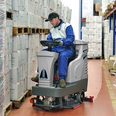 Homem a operar máquina de limpeza industrial cinzenta num armazém com caixas brancas