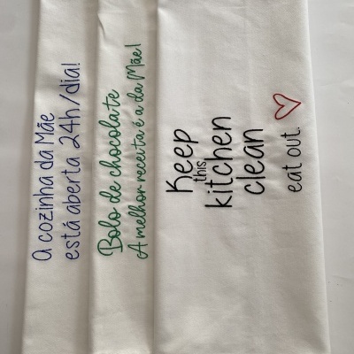 Três panos de cozinha brancos com frases bordadas coloridas dispostos lado a lado.