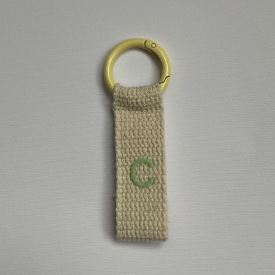 Porta-chaves com anel amarelo e tira de tecido bege com letra 'C' verde