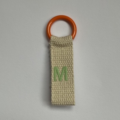 Porta-chaves com anel plástico laranja e tira de tecido bege com letra M verde