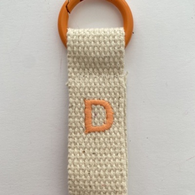 Porta-chaves com anel de plástico laranja e tira de tecido bege com letra D bordada em laranja