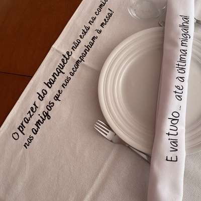 Toalha de mesa e guardanapo branco com texto bordado preto, prato branco, garfo e copo transparentes numa mesa de madeira