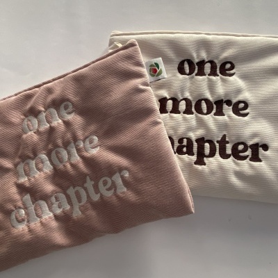 Duas bolsas de tecido bombazine rosa e bege com texto bordado 'one more chapter'.
