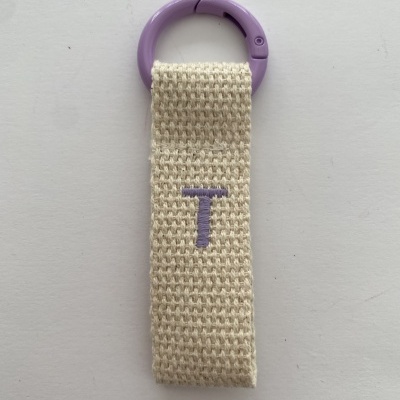 Chaveiro com argola de plástico lilás e tira de tecido bege com letra T bordada