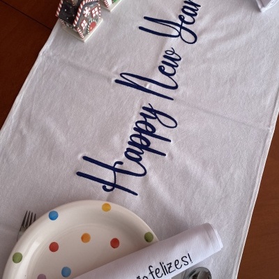 Toalha branca decorativa com texto 'Happy New Year' e pratos coloridos numa mesa de madeira