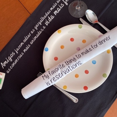 Mesa posta com prato branco de pontos coloridos, guardanapo branco com texto em inglês, copo de vidro e talheres sobre toalha preta com texto em português