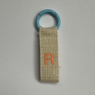 Chaveiro com argola azul clara e tira de tecido bege com letra R laranja