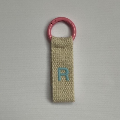 Chaveiro com argola plástica rosa e fita de tecido bege com letra R bordada.