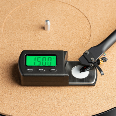 Medidor digital com display verde sobre superfície circular de cortiça e braço preto ajustado.