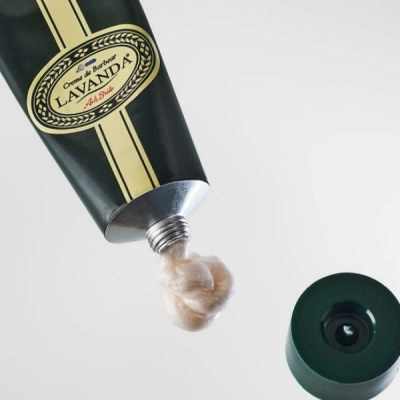 Tubo preto de creme de barbear Lavanda com creme bege saindo e tampa verde escuro ao lado
