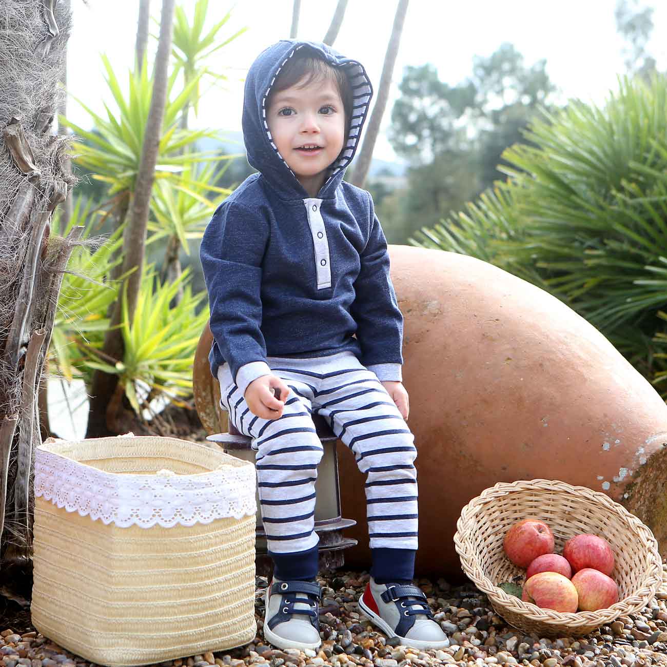Criança com roupa azul e branca sentada ao ar livre junto a cestos e plantas