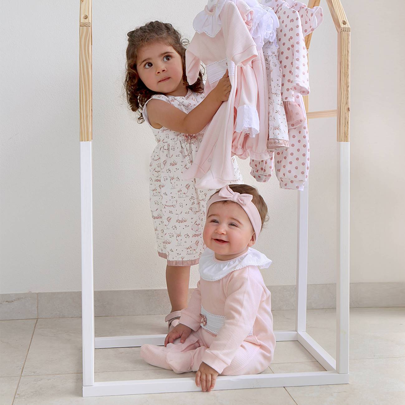 Duas crianças pequenas com roupa em tons rosa claro e branco junto a estrutura de madeira com roupa pendurada