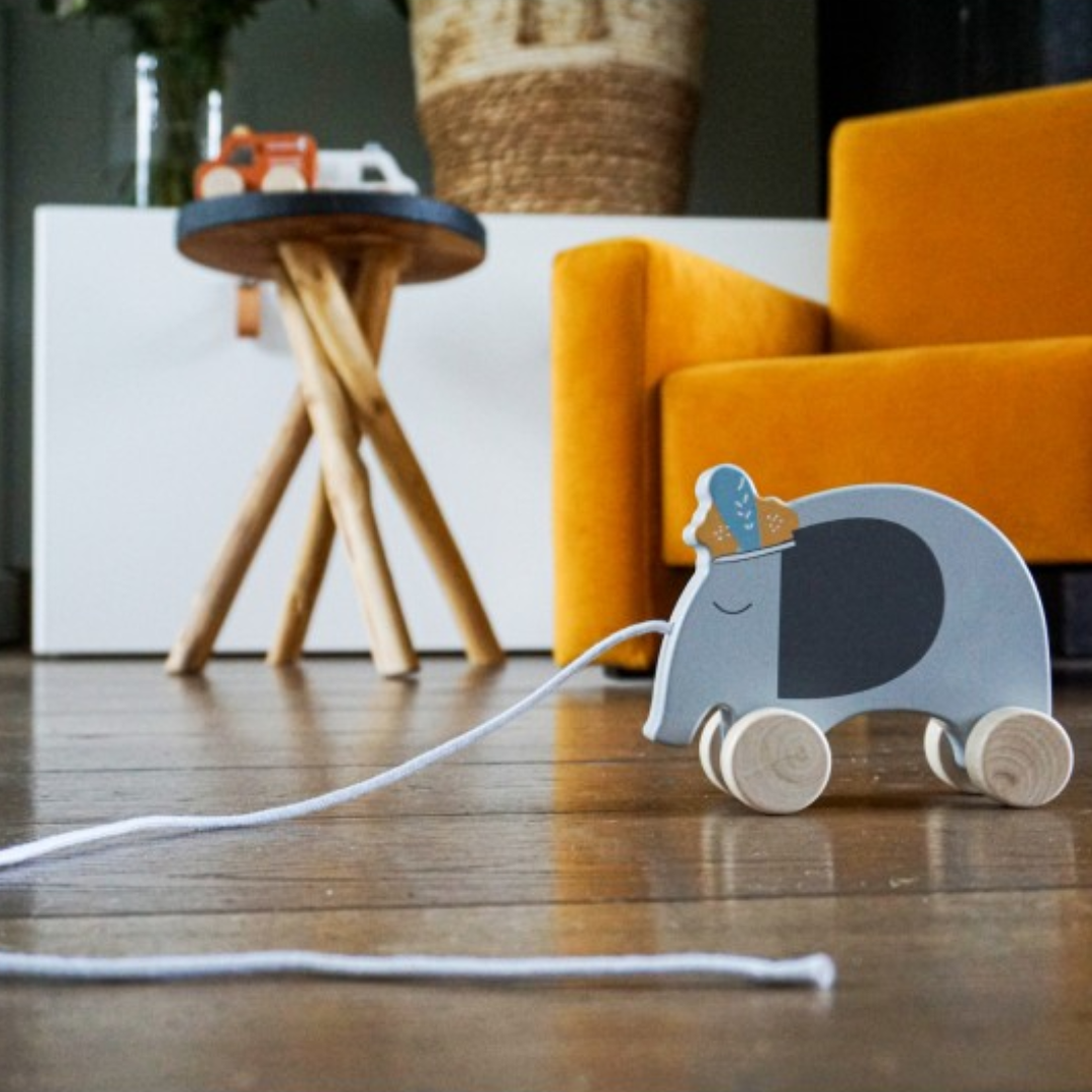 Brinquedo elefante de madeira com rodas e cordão, em chão de madeira junto a mesa e sofá amarelo.