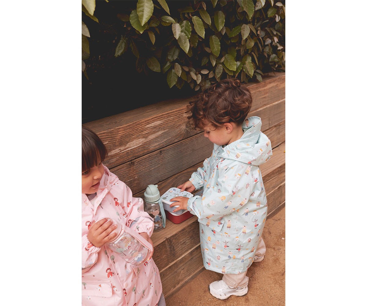 Duas crianças em capas de chuva coloridas, uma rosa e outra azul claro, perto de parede de madeira com plantas.