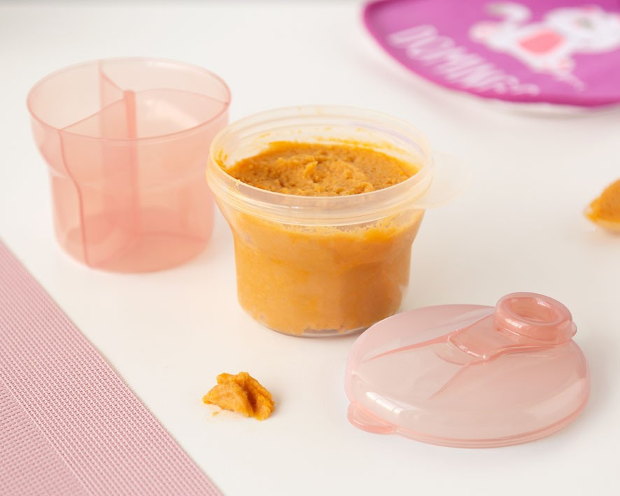 Recipiente transparente com tampa rosa contendo purê laranja, ao lado de copo rosa e prato rosa.