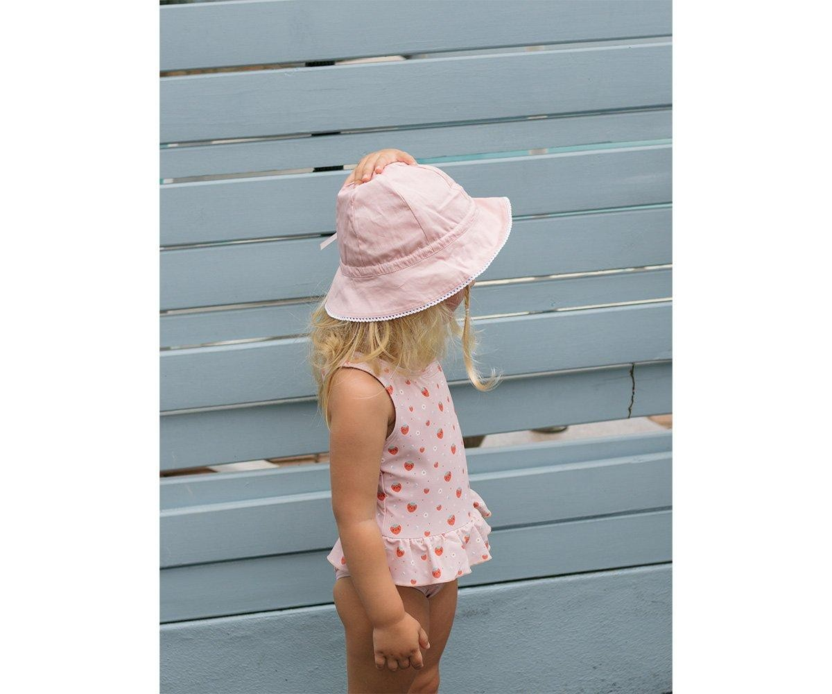 Criança de pé com chapéu rosa e maiô cor-de-rosa com corações vermelhos junto a vedação azul