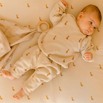 Bebé deitado em cama bege com padrão de girafas, vestido com roupa bege de girafas.