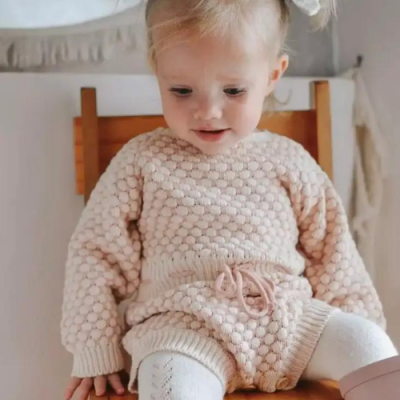 Criança sentada com roupa de malha bege com padrão de bolinhas, suéter e calções em malha