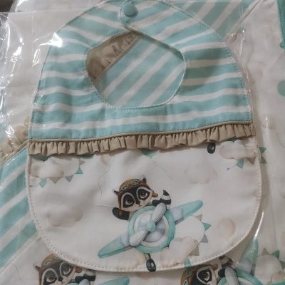 Babete de bebé com desenho de avião e guaxinim em tons verdes, brancos e bege