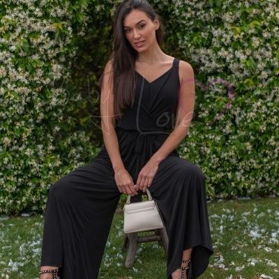 Mulher com macacão preto largo e mala bege sentada em banco exterior com arbustos floridos ao fundo