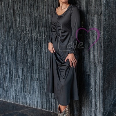 Mulher de vestido preto com detalhe franzido, botas prateadas, parede texturizada escura ao fundo