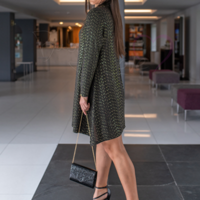 Mulher com vestido curto preto e dourado, salto alto preto e mala preta numa área interna moderna.